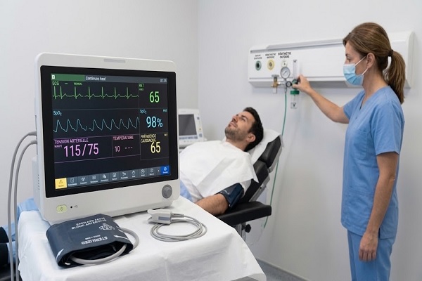 Un moniteur médical affichant des ondes ECG et SpO2 stables en temps réel pendant une chirurgie FUE dans une clinique espagnole. Une infirmière ajuste le débitmètre d'oxygène médical centralisé, démontrant la surveillance continue des signes vitaux et le matériel d'urgence dans les cliniques capillaires